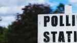 A sign reading 'polling station' in bold black letters, backed by trees and cloudy sky representing 2024 election.