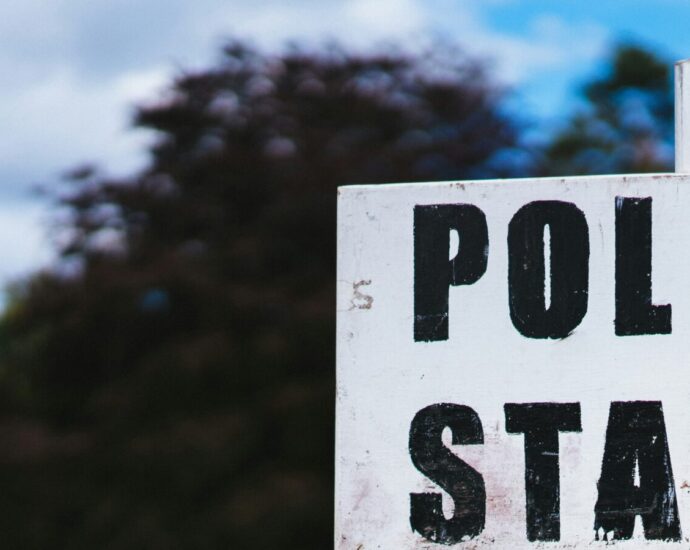 A sign reading 'polling station' in bold black letters, backed by trees and cloudy sky representing 2024 election.