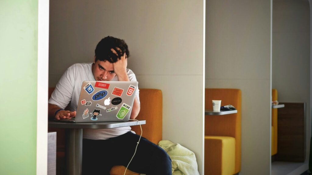 A young man sitting in a booth at his laptop covered in stickers with his head leaning on his hand during fresher's week