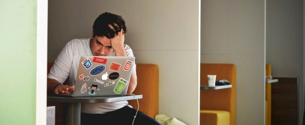 A young man sitting in a booth at his laptop covered in stickers with his head leaning on his hand during fresher's week