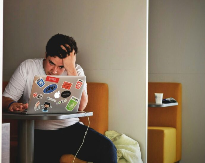 A young man sitting in a booth at his laptop covered in stickers with his head leaning on his hand during fresher's week