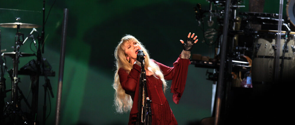 Stevie Nicks on stage in red dress singing into microphone with green lit background