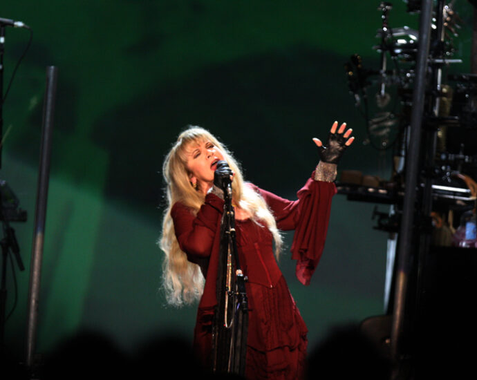 Stevie Nicks on stage in red dress singing into microphone with green lit background