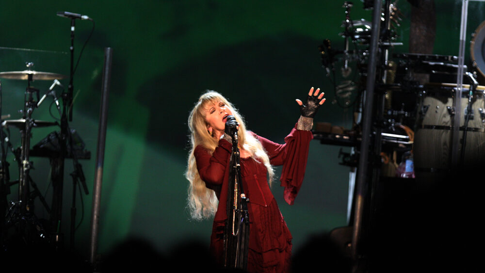 Stevie Nicks on stage in red dress singing into microphone with green lit background