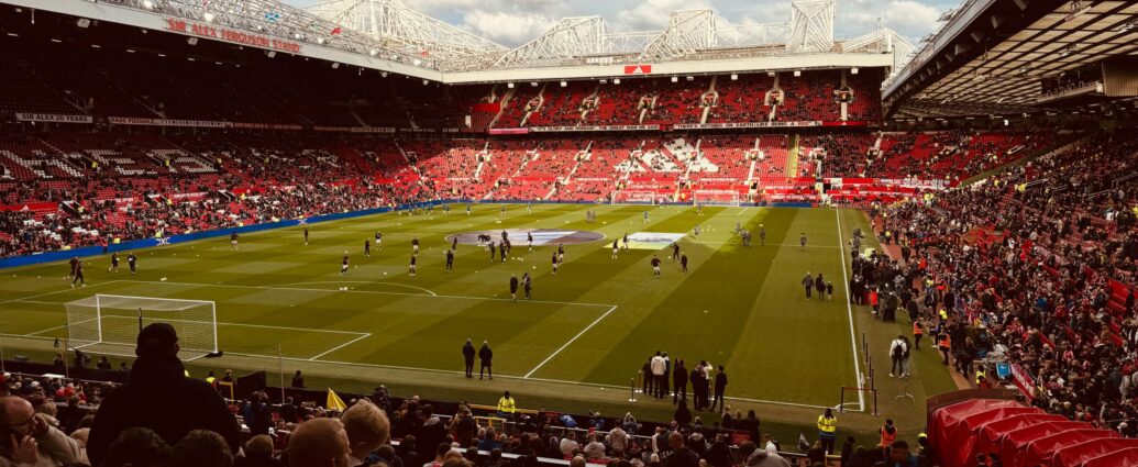 A football stadium with green pitch and red stand seating on bright day. Players on the pitch and fans in the crowd. [ten Hag]