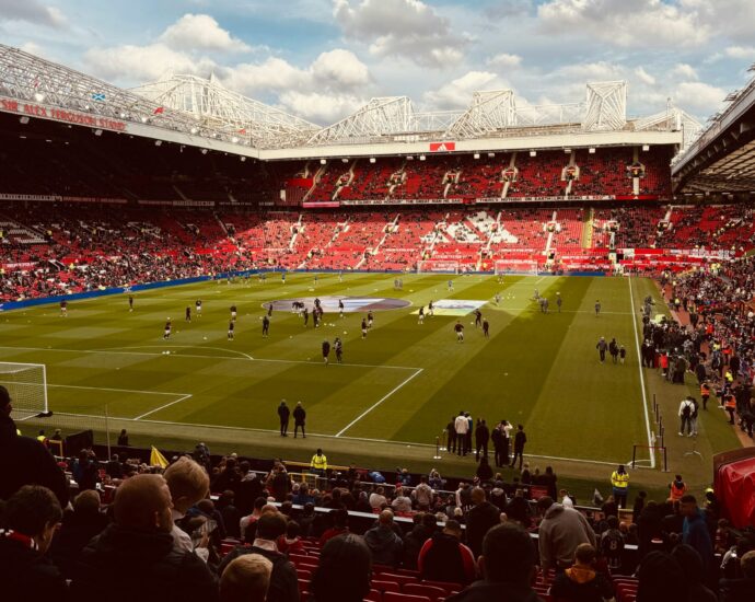 A football stadium with green pitch and red stand seating on bright day. Players on the pitch and fans in the crowd. [ten Hag]