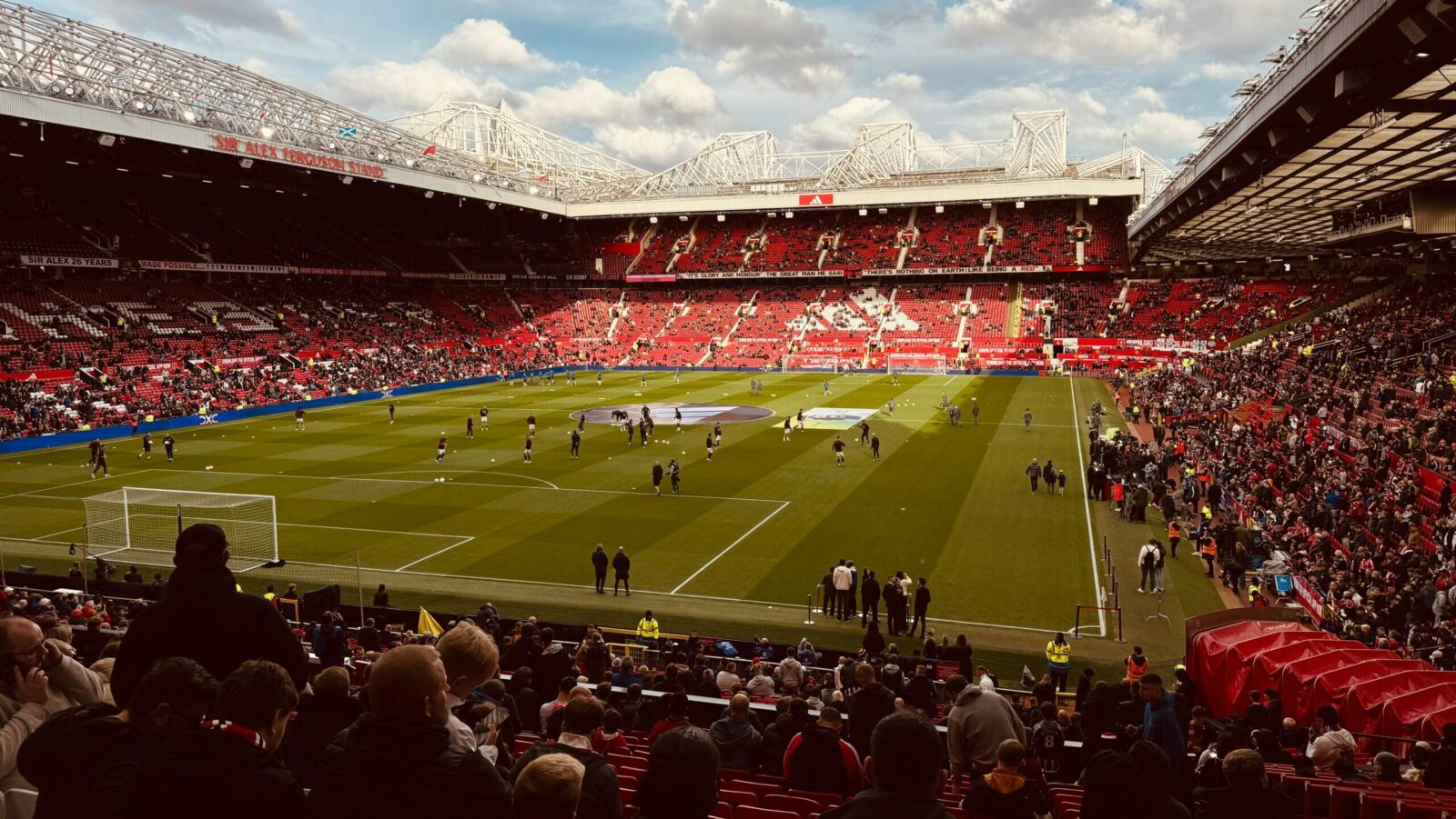 A football stadium with green pitch and red stand seating on bright day. Players on the pitch and fans in the crowd. [ten Hag]