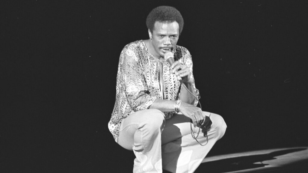 A black and white image of Quincy Jones crouched down on stage holding a microphone