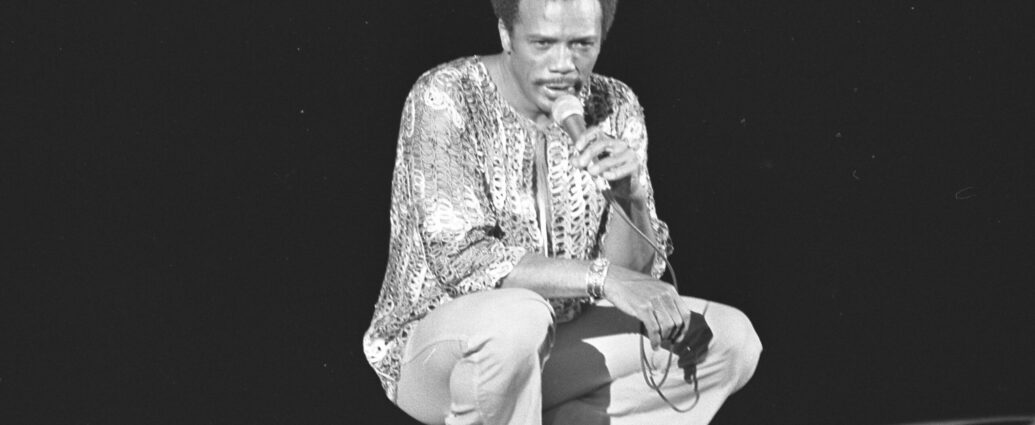 A black and white image of Quincy Jones crouched down on stage holding a microphone