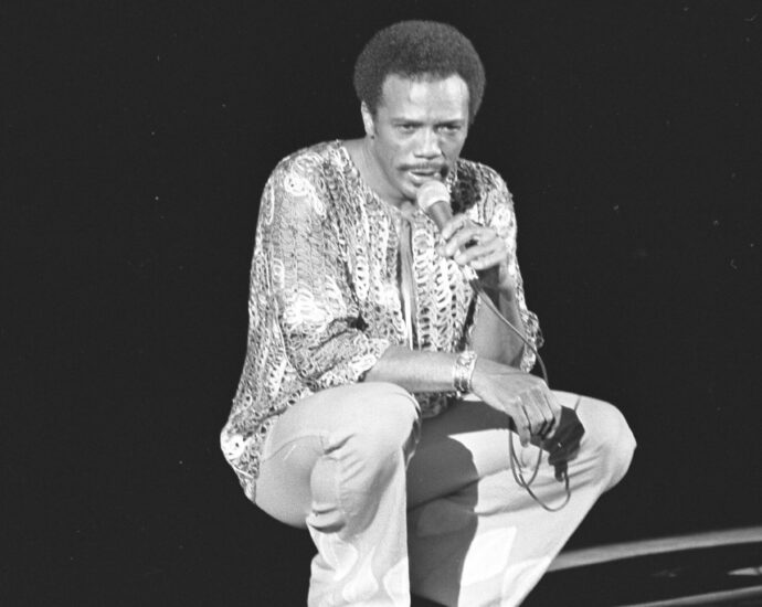 A black and white image of Quincy Jones crouched down on stage holding a microphone