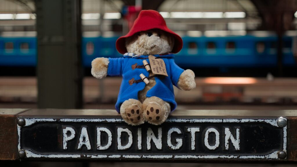 Paddington Bear in blue coat and red hat sitting on top of a sign reading 'Paddington'