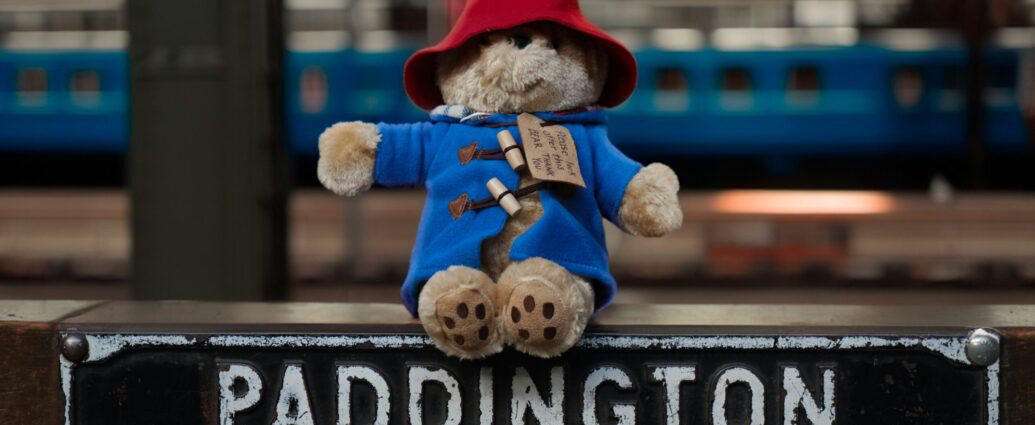 Paddington Bear in blue coat and red hat sitting on top of a sign reading 'Paddington'