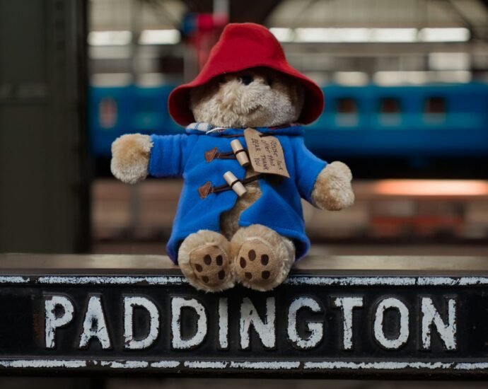 Paddington Bear in blue coat and red hat sitting on top of a sign reading 'Paddington'