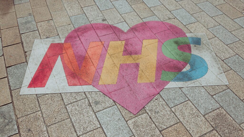 NHS written on pavement in red, yellow, green letters, backed by pink heart. NHS mental health funding.