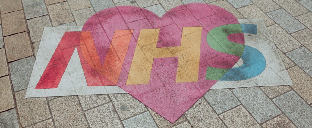 NHS written on pavement in red, yellow, green letters, backed by pink heart. NHS mental health funding.