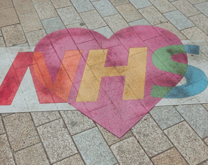 NHS written on pavement in red, yellow, green letters, backed by pink heart. NHS mental health funding.