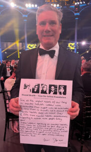 Kier Starmer in white shirt, black jacket and bow tie holding sign with black writing