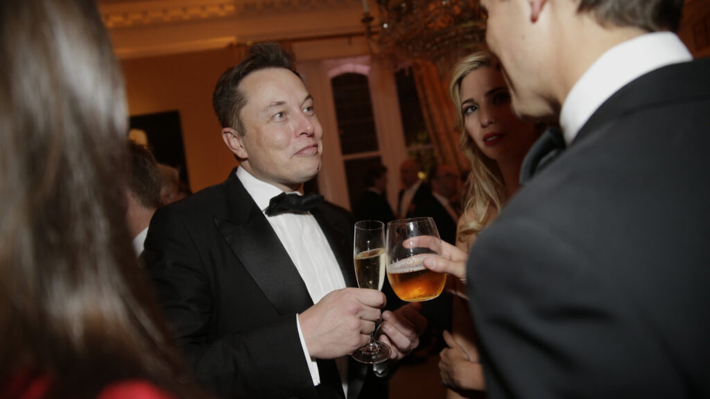 Photo of Elon Musk at an event, raising a drink towards another person