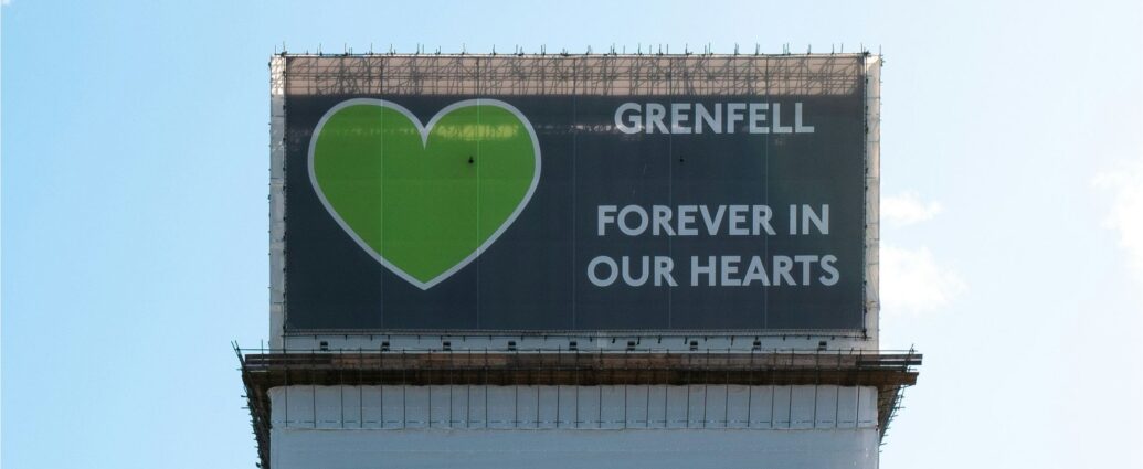 Grenfell tower stands against a blue sky, displaying the cover at the top of the tower which reads "Grenfell, forever in our hearts."