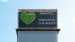 Grenfell tower stands against a blue sky, displaying the cover at the top of the tower which reads "Grenfell, forever in our hearts."