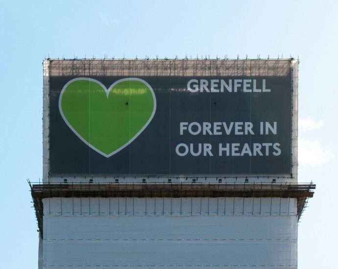 Grenfell tower stands against a blue sky, displaying the cover at the top of the tower which reads "Grenfell, forever in our hearts."