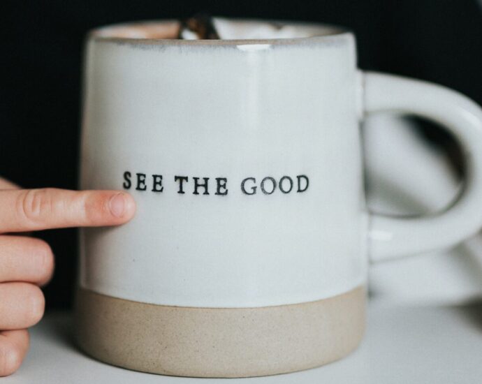 A finger is seen pointing at the words "see the good" engraved on a white mug.