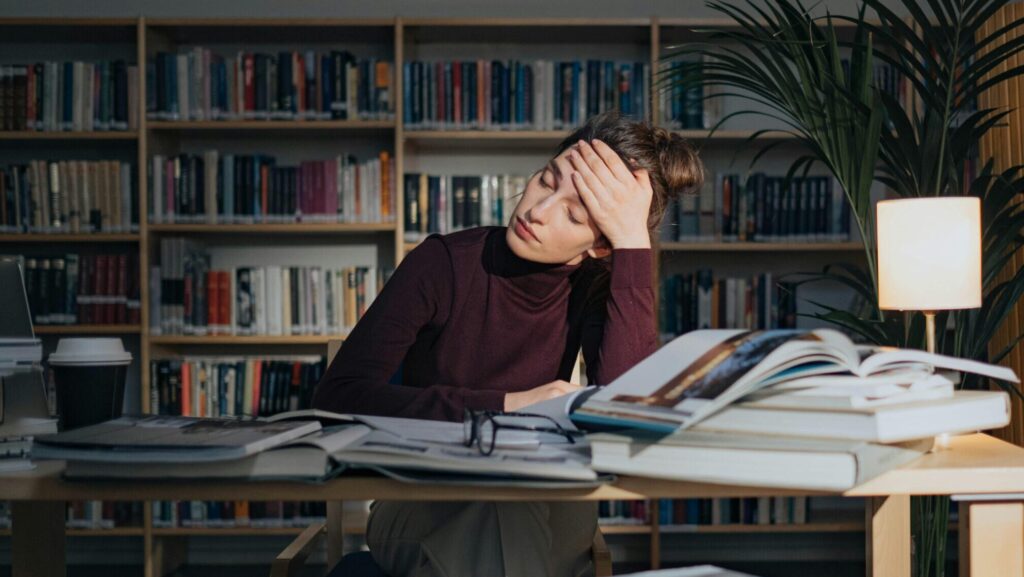 A person who appears stressed, with their head pressed against their hand, sitting at a table with books piled up. Featured photo for an article about student health and University Mental Health Day