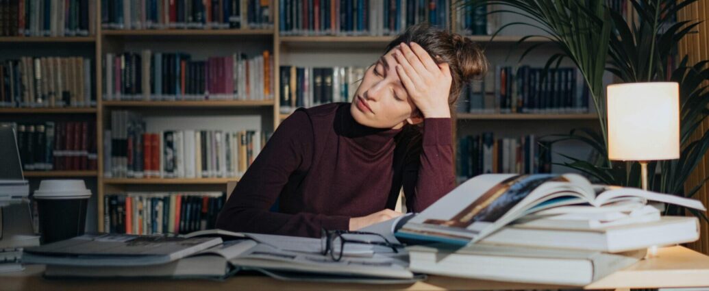 A person who appears stressed, with their head pressed against their hand, sitting at a table with books piled up. Featured photo for an article about student health and University Mental Health Day