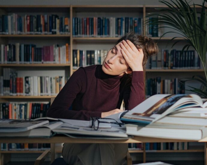 A person who appears stressed, with their head pressed against their hand, sitting at a table with books piled up. Featured photo for an article about student health and University Mental Health Day