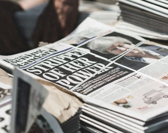 A stack of newspapers to represent the theme of 'journalism.' The image is blurred but the front page reads 'Stripped of her power' and shows Teresa May.