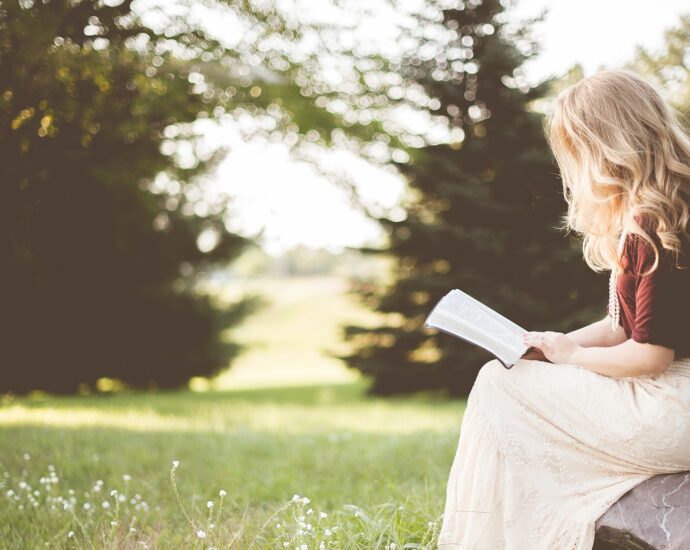 Girl sitting reading a book outside with grass and trees in the background potentially sunrise on the reaping